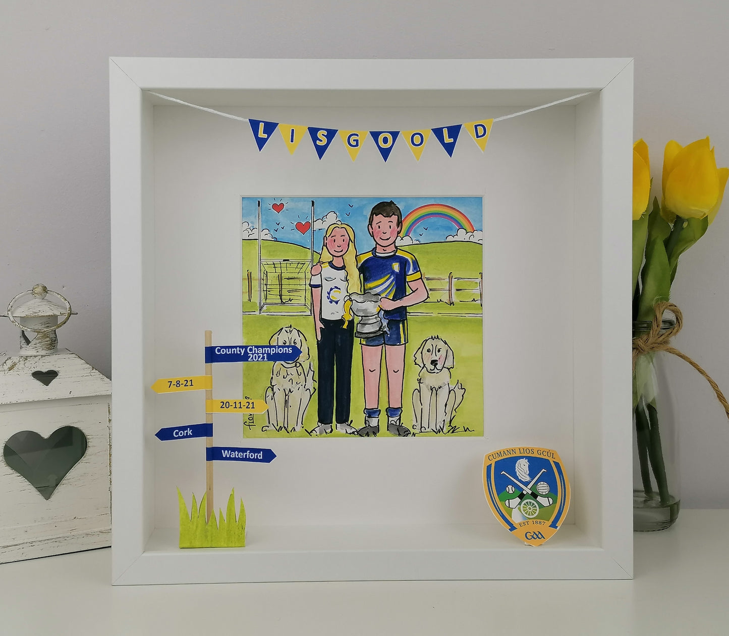 Framed artwork with a gaelic hurling couple and dogs, celebrating achievements, on a white surface with decorative items.