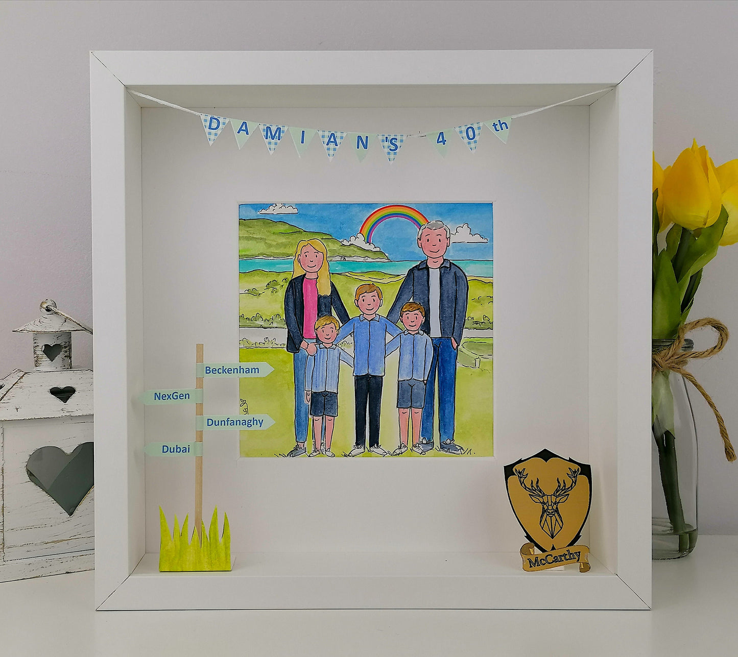 A colorful illustration of a family in a garden setting, with a banner that reads 'DAMIANS 40th', indicating a 40th birthday celebration. The artwork is framed in a white box frame.