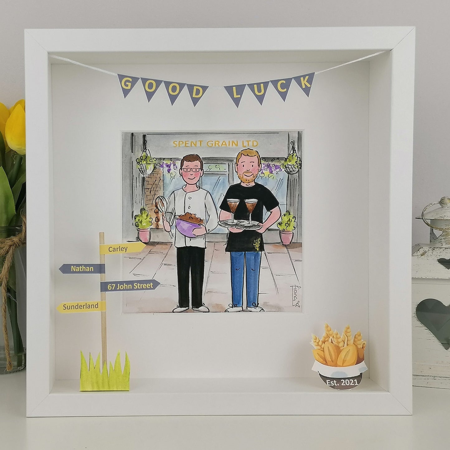 Framed artwork with two cartoon characters holding drinks, 'Good Luck' banner, and small cupcake decoration.