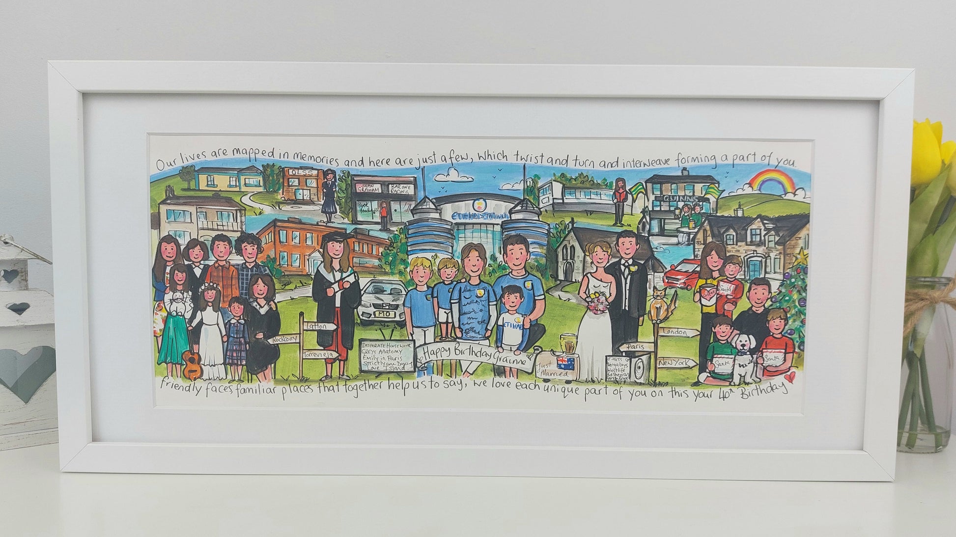Framed artwork of a wedding scene with people and buildings, on a white surface.