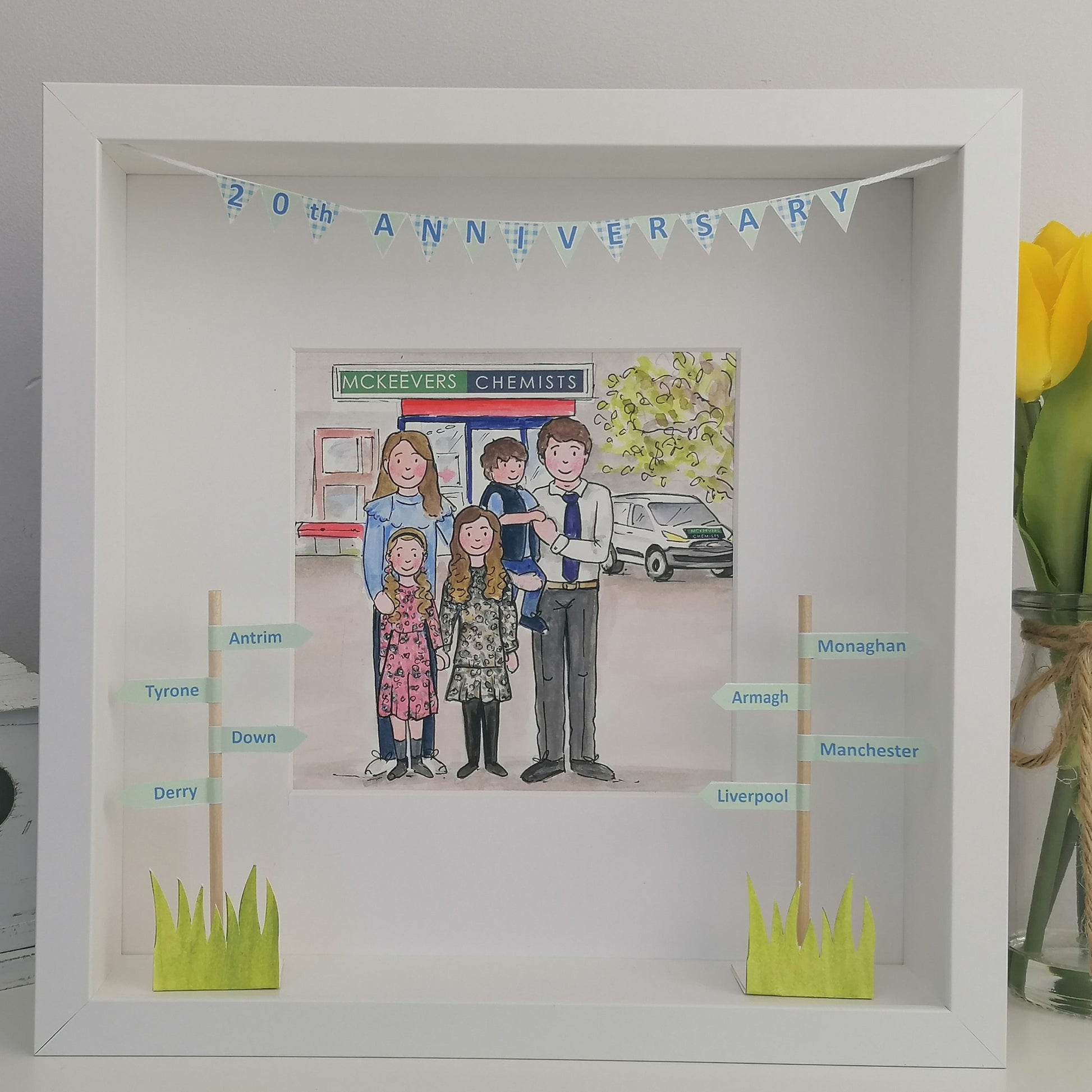 Shadow box with a family illustration and text, next to a vase of yellow tulips.