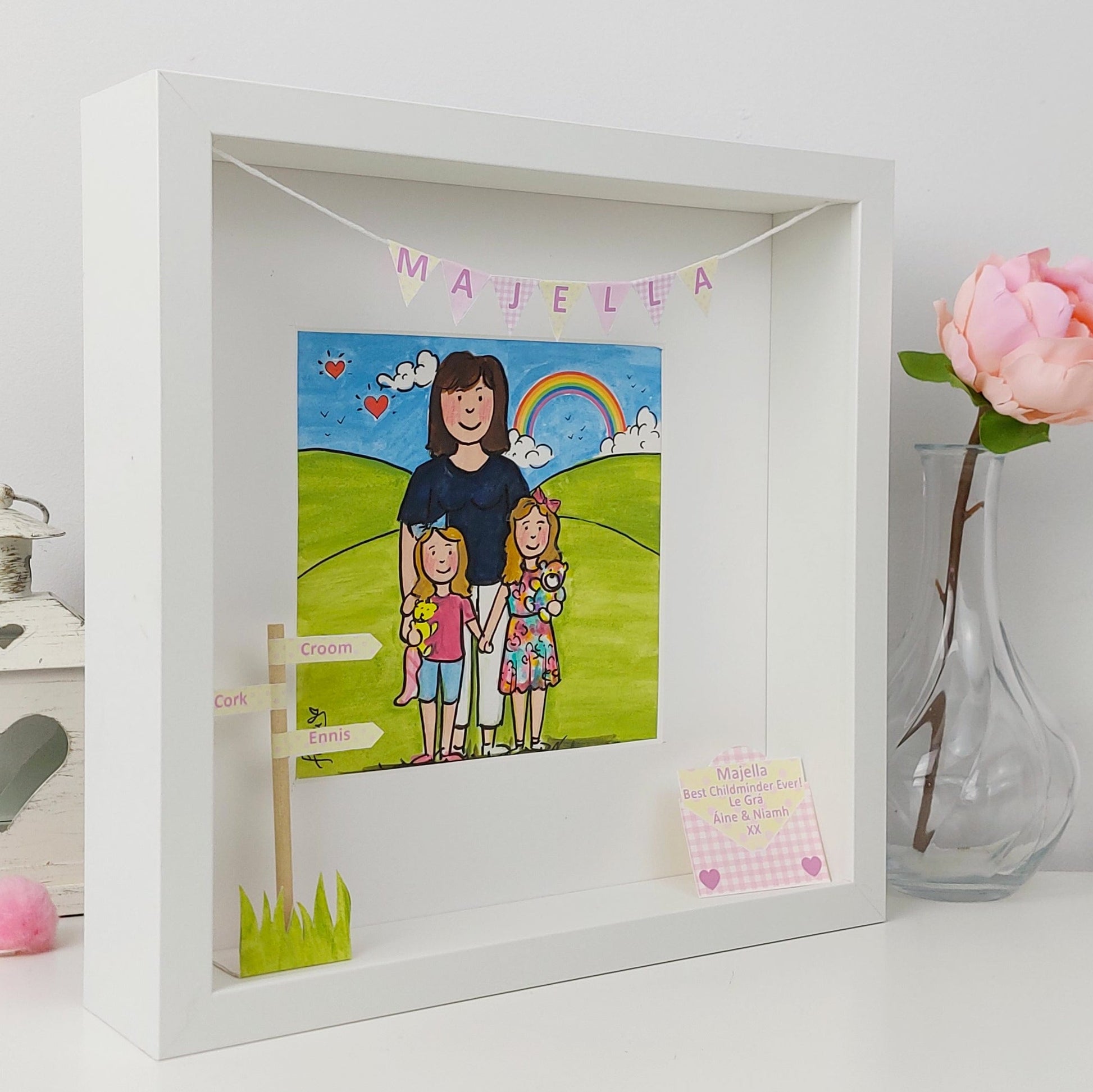 Framed illustration of a woman and two children with a rainbow, placed on a white surface with a vase and flower in the background.