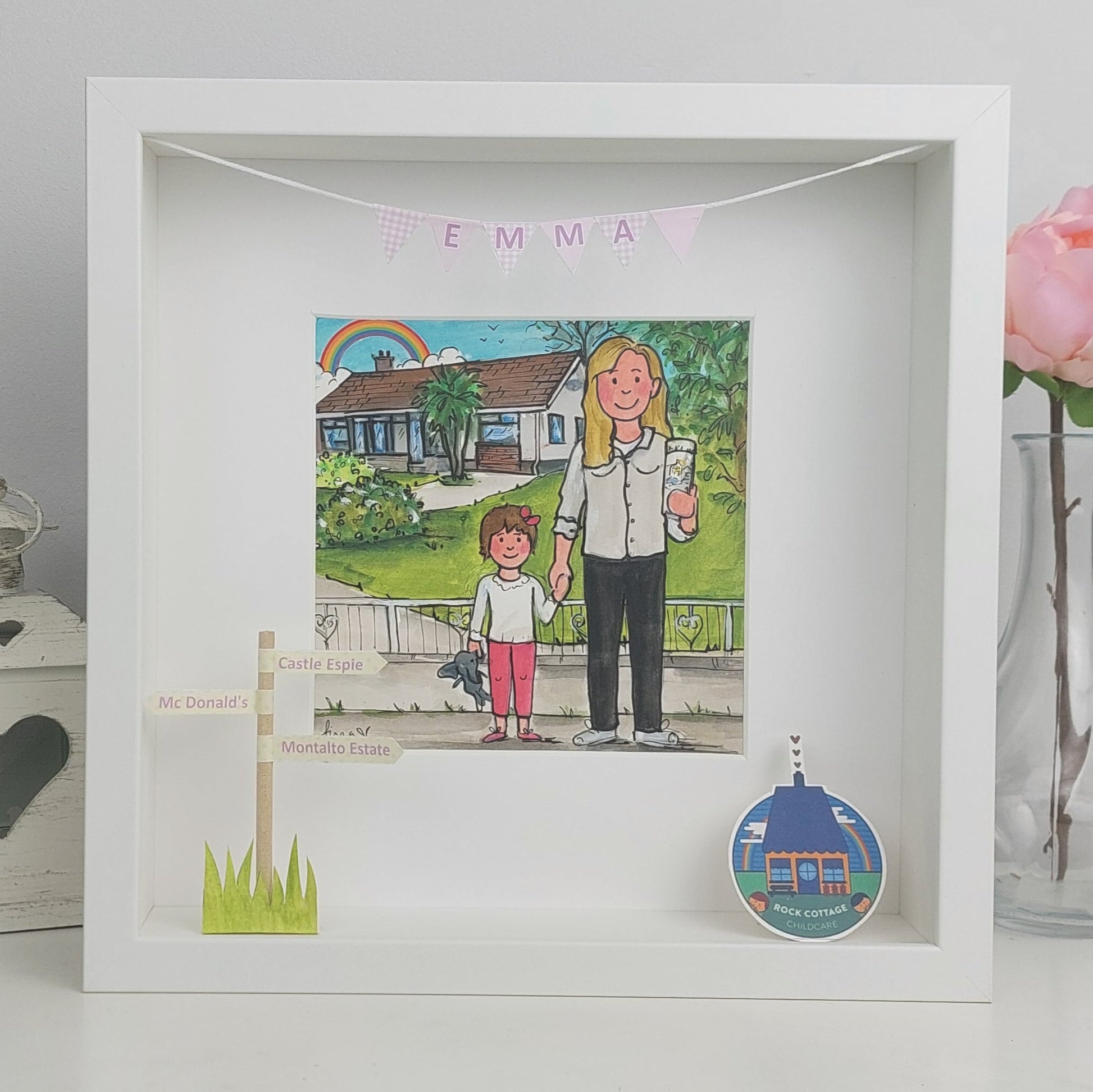 White framed picture with illustration of a woman and child, rainbow, and house, next to a decorative item.