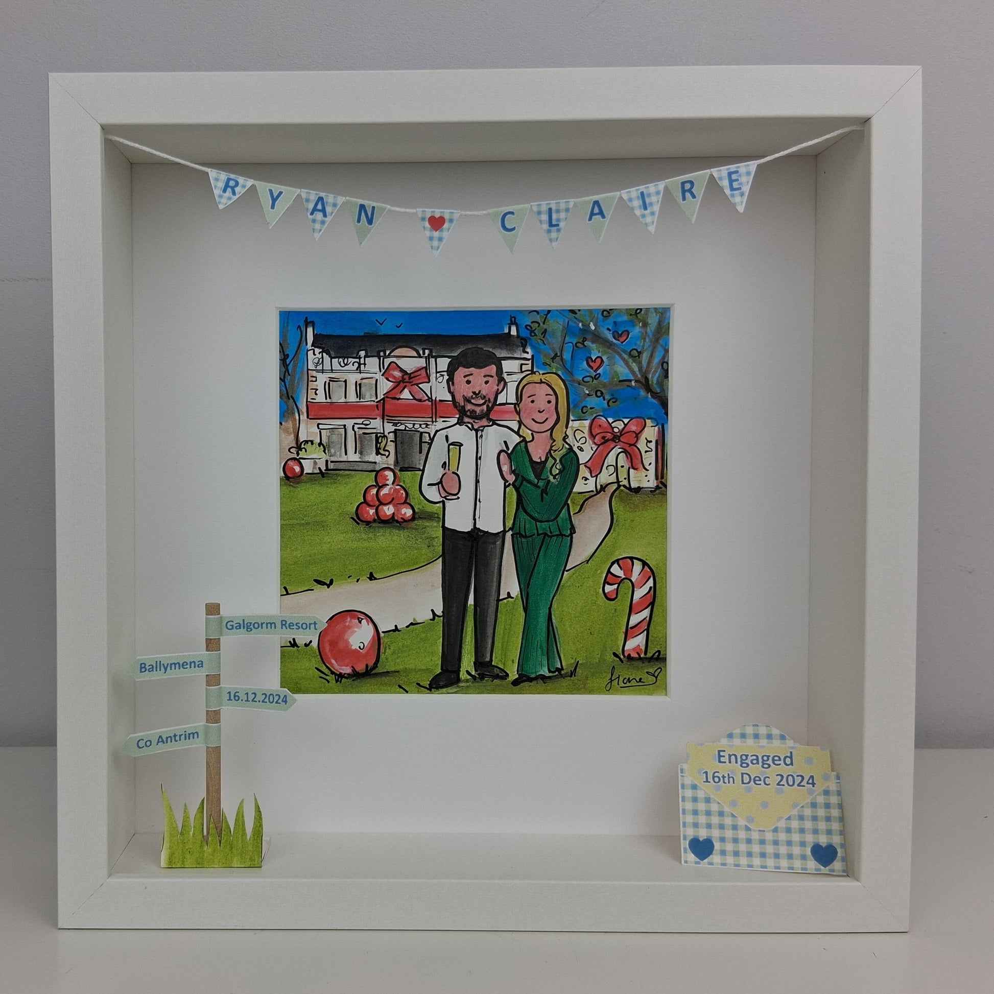 Framed artwork of an engaged couple at the galgorm with celebratory elements, including a house and candy canes, on a gray background.