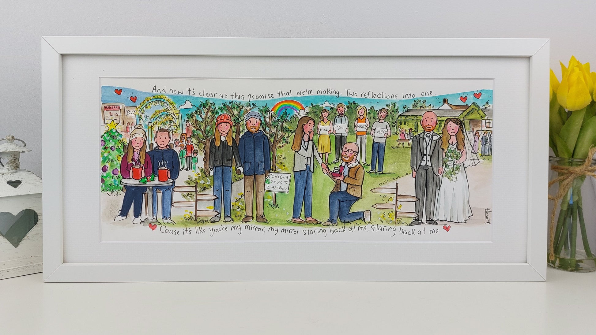 Framed artwork of a wedding scene with people and decorations, on a white surface with a vase of yellow flowers.