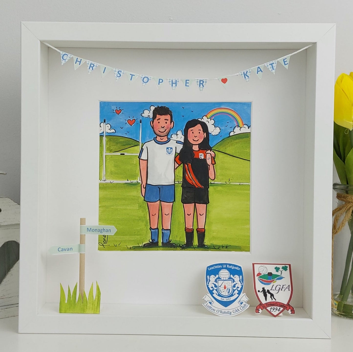 Framed artwork of a couple holding hands with a rainbow, surrounded by small flags and a vase of yellow roses.