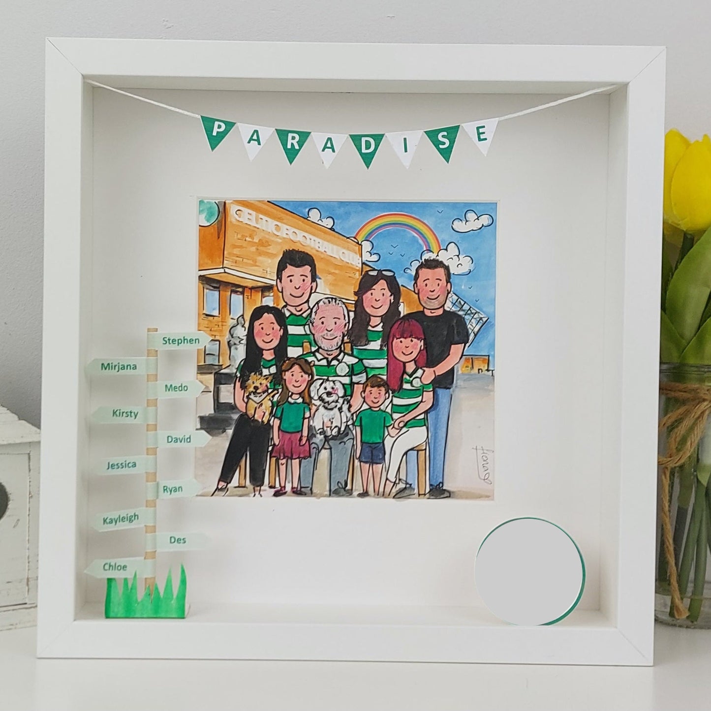 Framed artwork of a family with green accents, including a 'Paradise' banner and a family tree, on a white surface.