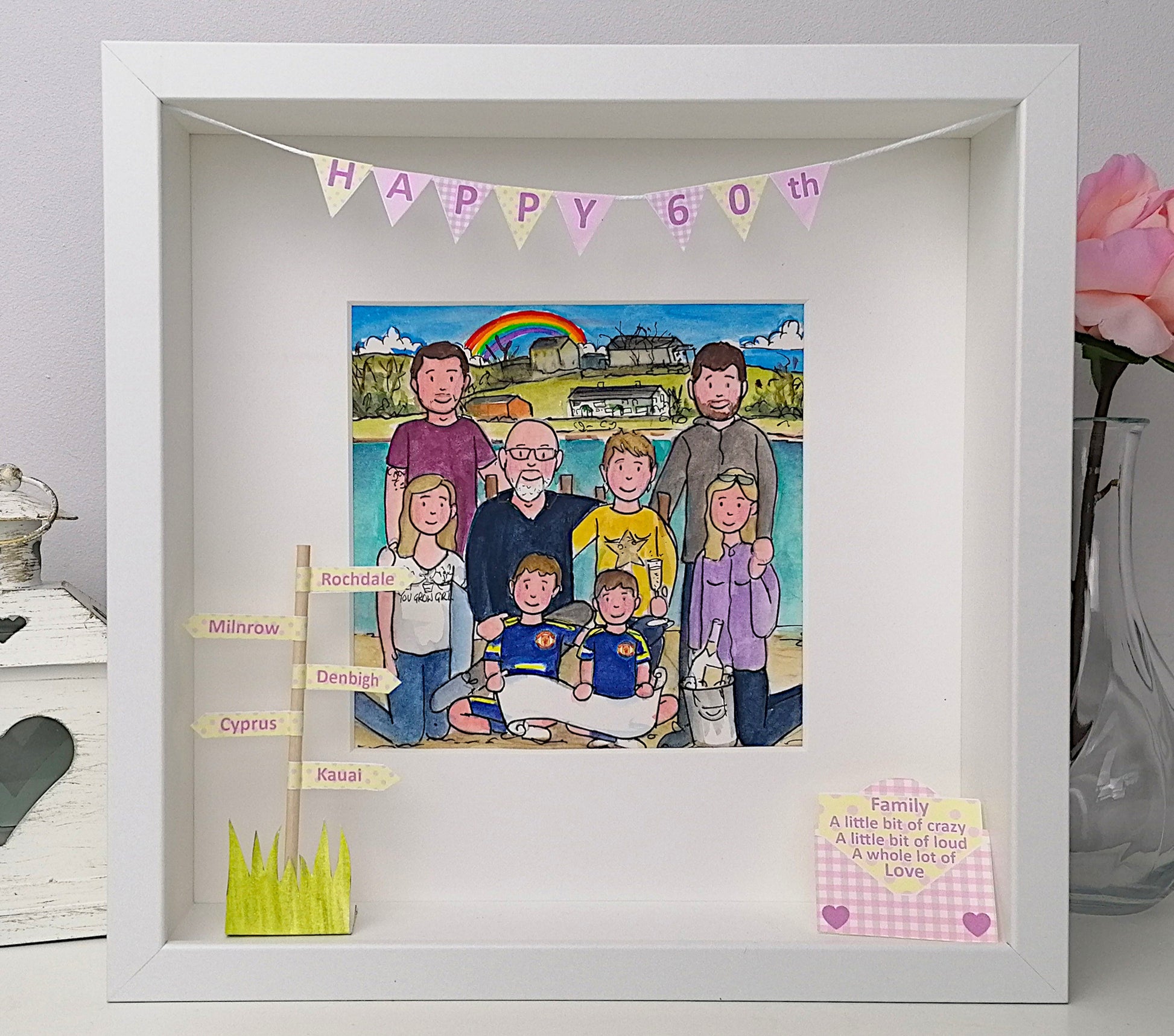 Framed artwork with a family portrait and 'Happy 60th' banner, placed on a surface with a vase and book in the background.