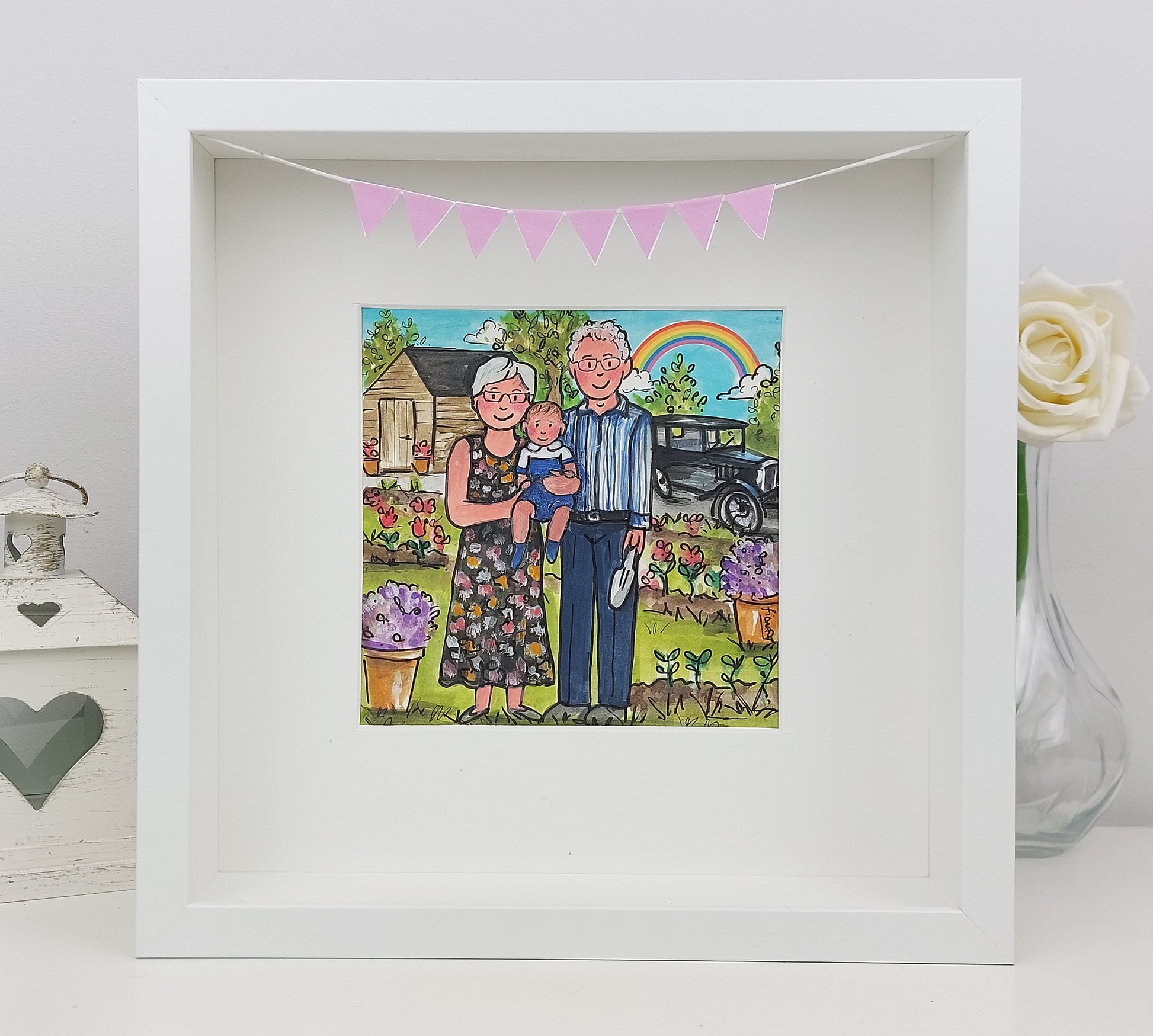 A framed artwork featuring a colorful illustration of a grandfather and grandmother with a cartoon-style garden and car in the background, surrounded by decorative pink bunting.