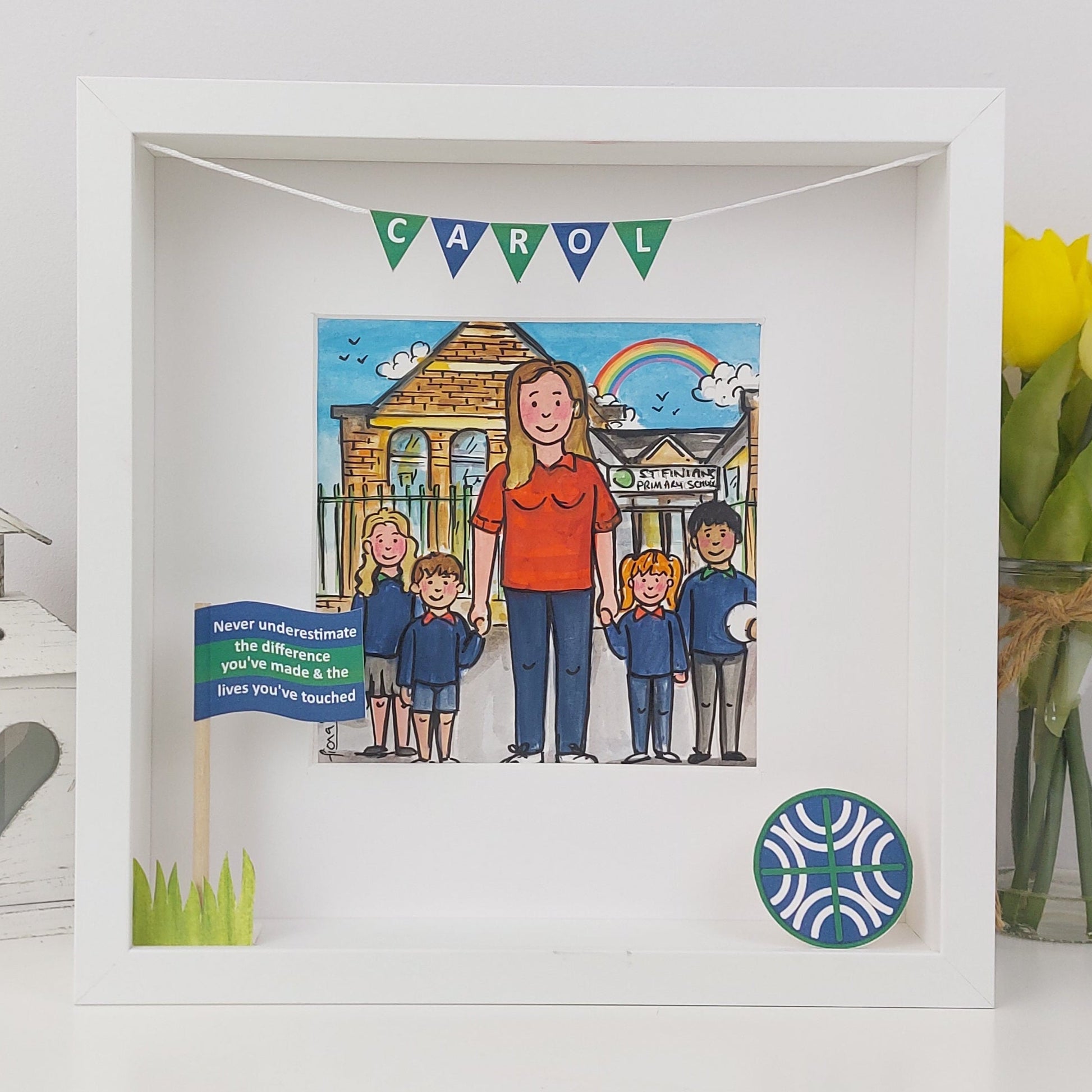 Framed artwork of a woman with four children in front of a house, with 'CAROL' above, on a white surface with flowers.