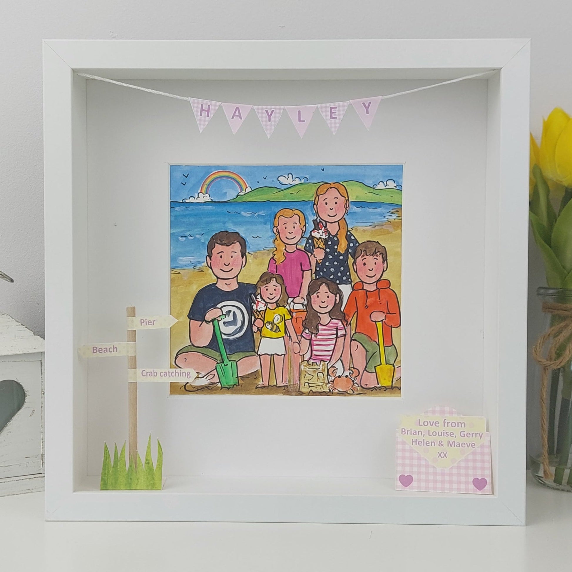 Framed illustration of a family on a beach with 'Hayley' above, next to a vase of tulips.