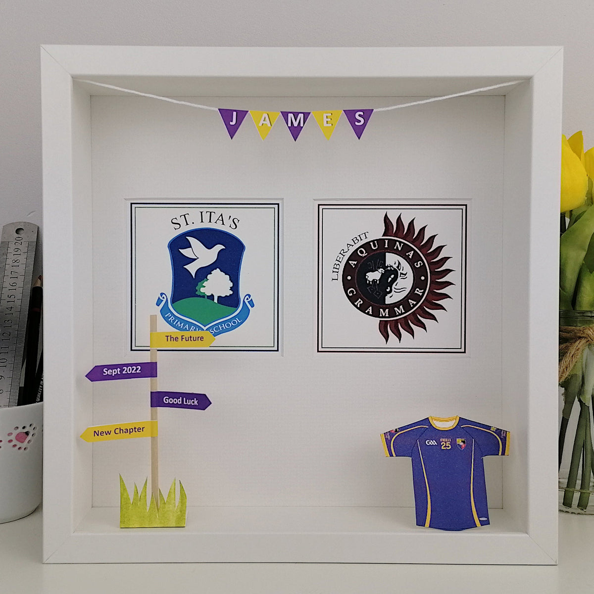 Decorative shadow box with school logos and a sports jersey, featuring a vase of yellow tulips on the right.