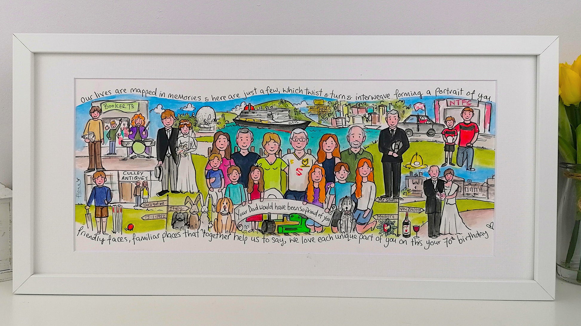 Framed artwork of a family portrait with various scenes and people, on a white surface with a vase of yellow flowers.