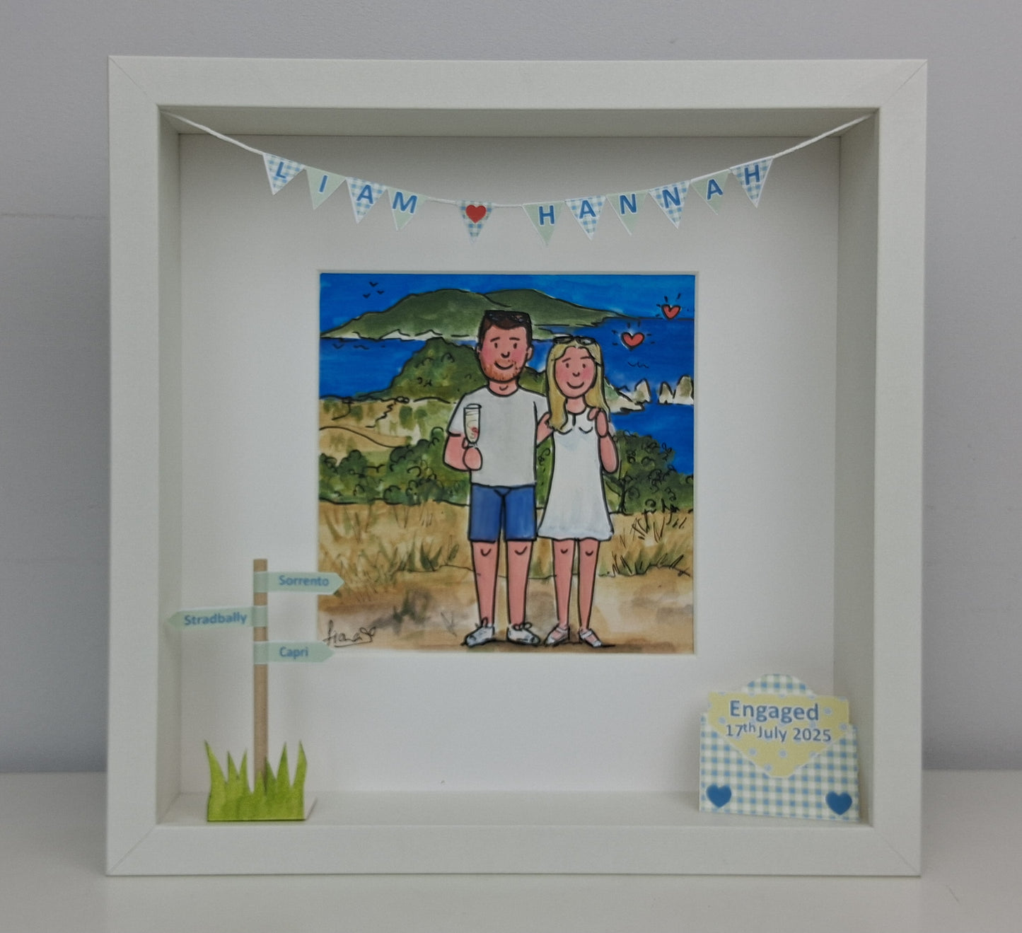 Framed artwork of an engegd couple on a beach with 'Liam Hannah' banner above, on a gray background.